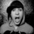 Black-and-white pixelated image of a person with mouth open, wearing a dark cap and light hooded garment, set against solid orange background with pixelated text.