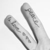 Two fingers with text and symbols tattooed on them, one finger reads 'GLORY' with heart symbols and the other reads 'PEACE' with a checkmark symbol.