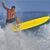 Pixelated image of a person in mid-air above water, holding a yellow surfboard with one hand, wearing a brown shirt and dark shorts, against a blue sky background.