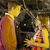 Two figures in a close embrace, one in a yellow top and pink skirt, the other in a yellow dress with red accents, against a dark textured background.