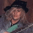 Pixelated static image of a person wearing a dark hat and light-colored clothing with green accents, set against a blurred background.