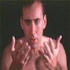 A shirtless man with short brown hair stands against a dark background, holding both hands up near his chest with palms facing forward.