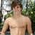 A pixelated static image shows a shirtless person with brown hair standing outdoors against a blurred green and blue background.