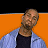 Pixelated image of a person with short brown hair wearing a dark shirt and light blue jacket against an orange background.