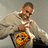 A person in a light gray hoodie with an orange cartoon face graphic points at the camera against a plain background, with repeated text elements on adjacent