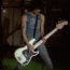 A person in a sleeveless denim shirt and blue jeans plays a white electric guitar with black pickguard on a dark stage with green lighting.