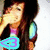 A person with long dark hair covers their mouth with one hand, wearing a colorful striped shirt with blue and green shoulder accents, and a heart-shaped icon in the corner.