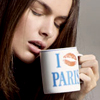 A person with long brown hair holds a white mug with blue text and an orange lip print, drinking from it against a gray background.