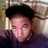 Pixelated static image of a person with dark hair wearing a black cap and dark clothing against a blurred background.