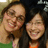 Two people with brown hair smiling closely together, one in green and orange clothing against a dark background.