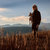 Pixelated static image of a person standing on a grassy hillside facing left, with a hazy sky and distant horizon in the background.