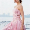 A person in a pink striped strapless dress stands against a blurred waterfront background with distant buildings.