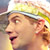 Pixelated close-up of a person wearing a light-colored hat with green and yellow accents, visible facial features including eyes, nose, mouth, and ear, against a blurred background.