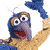 Pixelated blue puppet with large eyes, red mouth, and brown arms holding a blue object above its head against a white background.