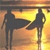 Silhouetted figures of two people standing on a beach at sunset with one figure extending an arm toward the other against a gradient sky and water background.