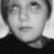 Static grayscale image of a person's face with short hair, wearing a dark top, against a light background.