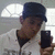 A person wearing a dark blue cap and white zip-up jacket holds a dark smartphone, with a bright arched window in the background.