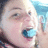 Pixelated close-up of a person's face with light skin tone, dark hair, and a blue object near the mouth, set against a light background with blue and brown tones.