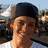 Pixelated portrait of a person wearing dark cap and white shirt against blurred outdoor background with text elements below.