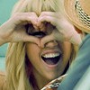 A person with long blonde hair forms a heart shape with both hands over their face, mouth open in a wide smile, wearing a light blue garment and a straw hat with a decorative band.