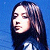 Pixelated portrait of a person with long dark hair against a blue background, wearing a dark top with light accents.