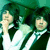 Three figures in white shirts and dark jackets stand against a green background with pixelated textures.