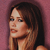 Pixelated close-up of a person with long brown hair against a pinkish-purple background, showing partial facial features and shoulder area.