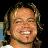 A pixelated close-up of a person with light brown hair smiling broadly, showing teeth, against a dark background with blurred light-colored elements.