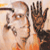 Pixelated static image shows a person with light skin holding a dark object near their face, another hand raised in the foreground, and an orange garment against a pale background.