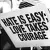 Black-and-white static image of a sign with bold text reading HATE IS EASY; LOVE TAKES COURAGE in three lines.