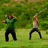 Two figures in green and beige shirts perform synchronized swinging motions with a long object against a grassy background.