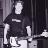 Pixelated grayscale image of a person in dark clothing holding an electric guitar, standing against vertical striped background.