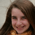 A young woman with long brown hair and a yellow top smiles at the camera in a static image.
