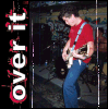 A person in a red shirt and blue jeans plays an electric guitar on stage, with vertical text reading "over it" along the left edge.