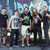 Group of seven figures in casual attire standing closely together against a blurred background with blue and gray tones.
