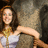 A woman in a white strapless dress with gold accents poses against a textured stone background, smiling with arms extended.