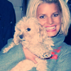 A person with short blonde hair holds a small white dog with curly fur, both facing forward against a dark background.