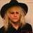 Pixelated portrait of a person with long blonde hair wearing a black wide-brimmed hat and dark clothing against a light beige background.