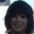 A person with dark shoulder-length hair and bangs smiles while making a hand gesture, wearing an orange garment against a light background.