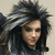 Pixelated close-up of a person with dark tousled hair, wearing a dark hooded garment with light gray accents, against a neutral background.