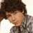 Pixelated close-up of a person with dark hair wearing a gray patterned shirt against a light background.