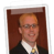 Static portrait of a person with light brown hair wearing glasses, dark jacket, white shirt, and patterned tie against reddish-brown background.