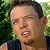 Pixelated close-up of a person with short dark hair, light skin, and a dark sleeveless top against a green foliage background.