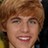 Pixelated close-up of a person with light brown hair, fair skin, and visible teeth in a neutral expression.
