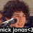 Pixelated image of a person with dark curly hair holding a microphone, wearing a dark shirt against a muted background.