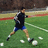 A person in a black shirt and blue shorts kicks a white soccer ball on a green field with scattered white markers.