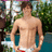 Pixelated image of a shirtless person standing outdoors with green foliage and blue water in the background.