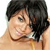 Pixelated close-up of a person with dark hair, light skin, and a neutral expression, wearing a light-colored top with a dark strap visible on the shoulder.