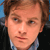 Pixelated close-up of a person with light brown hair, fair skin, and a neutral expression wearing a light blue collared shirt under a dark jacket against a blue background.