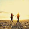 Two silhouetted figures holding hands on a beach at sunset, with warm yellow and orange tones dominating the sky and water.